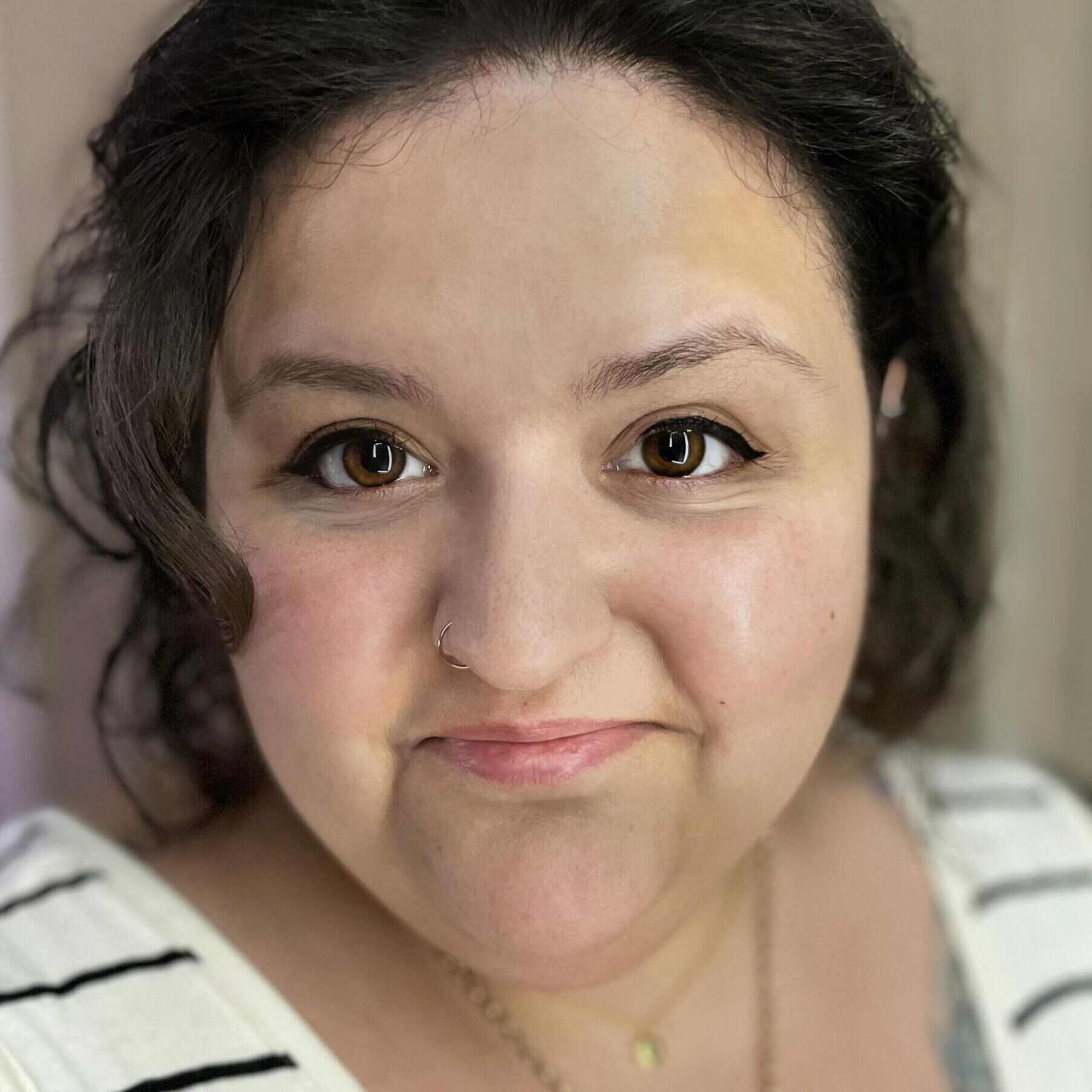 Cropped portrait of an India/white British woman with a closed-mouth smile, with short, curly, dark brown hair, brown eyes, and winged black eyeliner. Wearing a pale cream with thin horizontal black striped vest top, two gold necklaces of different lengths, and a tattoo of a bee on the left side of her chest peaking out from the top.