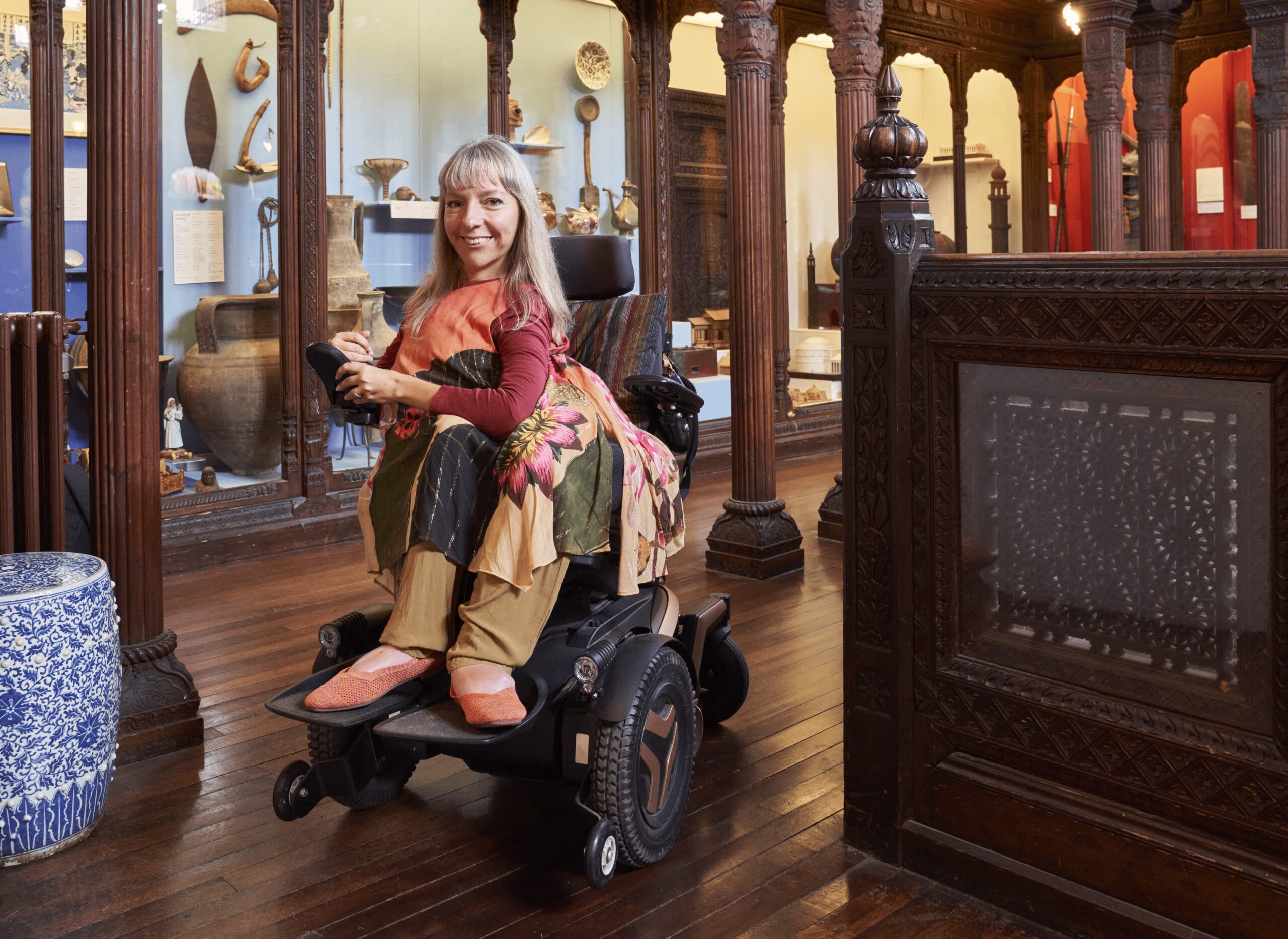 A full length photo of a white woman in her late 40’s with shoulder length silver blonde hair. She is a wheelchair user, and is smiling at the camera. She is in a museum setting, with dark wood columns and cabinets showing historic objects visible behind her.