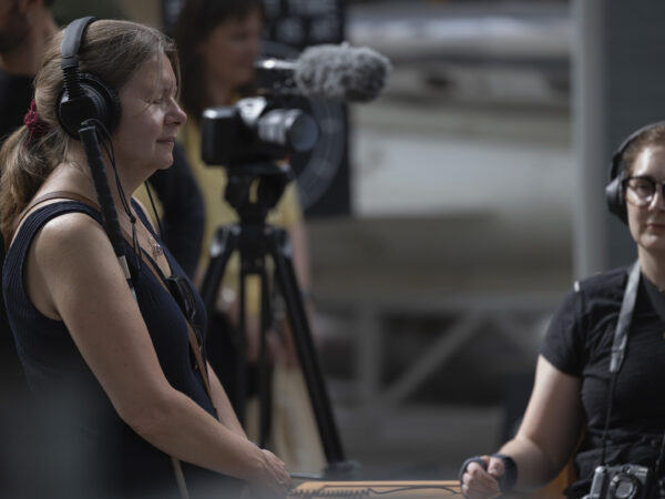 The photo shows Hannah standing the left, wearing headphones, eyes closed with an expression of focused, enjoyable listening. Her hair is pulled back in a red scrunchie, and she is holding her hands together in front. To the right, out of focus, is Charlotte seated. Also wearing large headphones, she appears to be looking into middle distance, to listen. A camera on a tripod with a fluffy microphone is behind and between them.