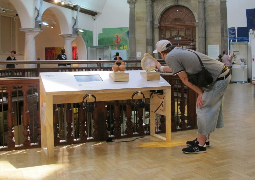 A photo of a multisensory exhibit. A man stands to the side of a table-top display in the Museum of Archaeology and Anthropology. He is bent over to get a closer look at a cross-section tactile of a Moche pot, which he has lifted to move the water inside.