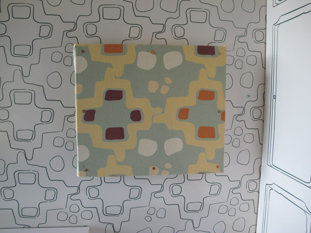 A photo showing a section of a museum panel. A square of brightly patterned wall fabric is pinned to a wall featuring the same pattern but in black and white.