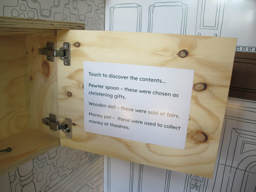 A photo of the inside of an open cupboard door. The door has a large text label which reads: Touch to discover the contents...
Pewter spoon - these were chosen as christening gifts.
Wooden doll - these were sold at fairs.
Money pot - these were used to collect money at theatres.