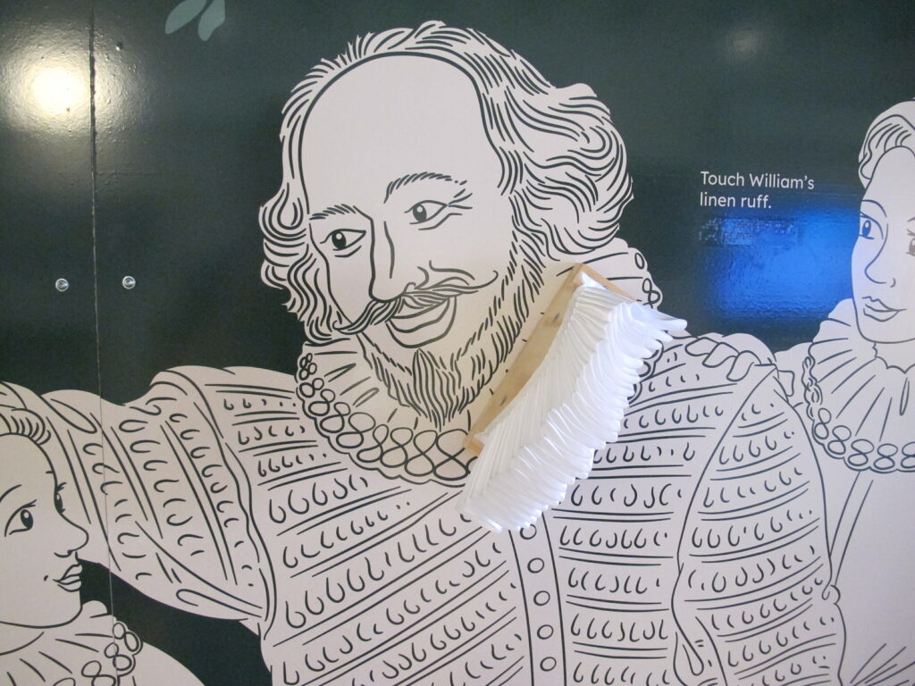 A photo showing a section of a museum panel. A green and white illustration of William Shakespeare is shown on a dark green background. Shakespeare is wearing Elizabethan clothes, half of his ruff is made out of real fabric. Text to the right invites you to touch the ruff. This is repeated in braille below.