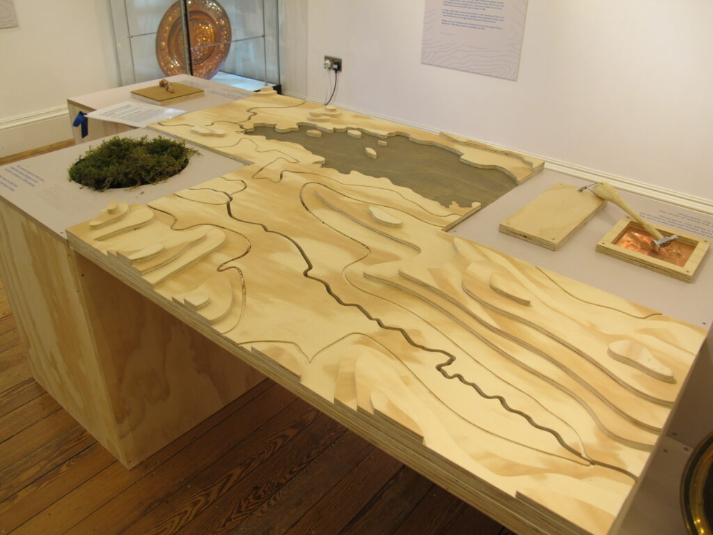 A photograph of the KSIA installation at Keswick Museum. Two large tactile wooden relief maps sit on a wooden table alongside some live moss and metal nuggets and hammer for visitors to touch.